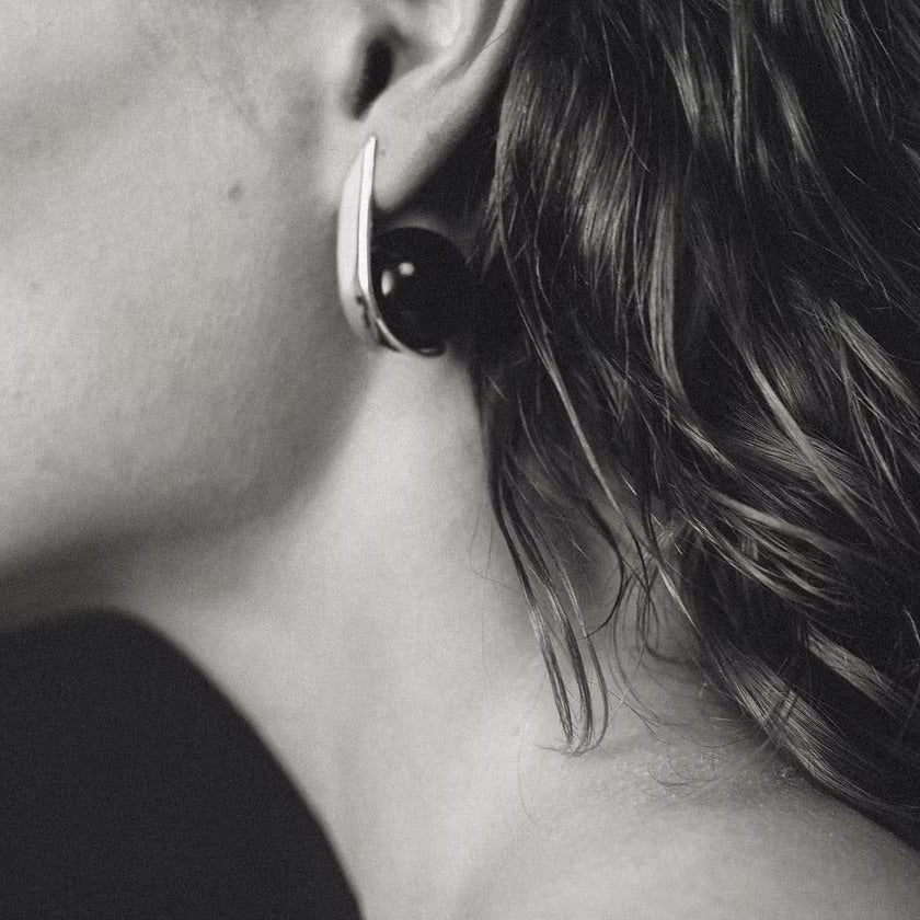 A close-up black and white photograph of a person's ear wearing a modern, curved earring with a dark circular amber and a silver-toned outer edge.