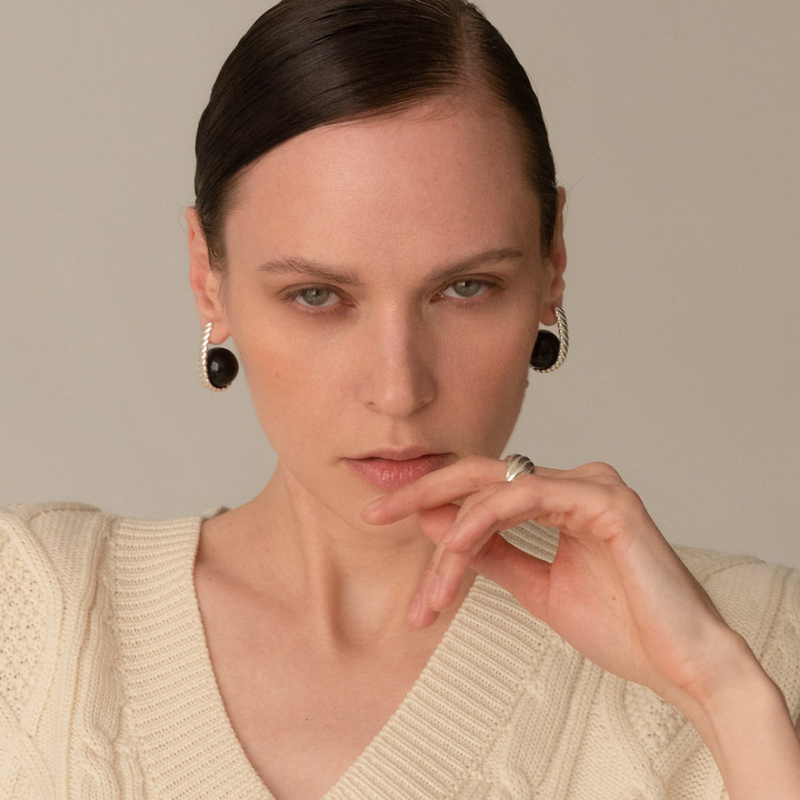 A close-up portrait of a woman wearing black oval drop earrings with silver-toned bezel settings and a silver ring on her finger, against a plain beige background.
