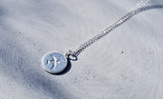 A silver pendant necklace with a circular disc featuring a stylized bird and star design, resting on a textured gray concrete surface.