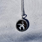 A silver pendant necklace featuring a bird and star design on a dark circular background, resting on a textured gray concrete surface.