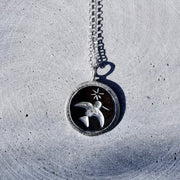 A silver pendant necklace featuring a bird and star design on a dark circular background, resting on a textured gray concrete surface.