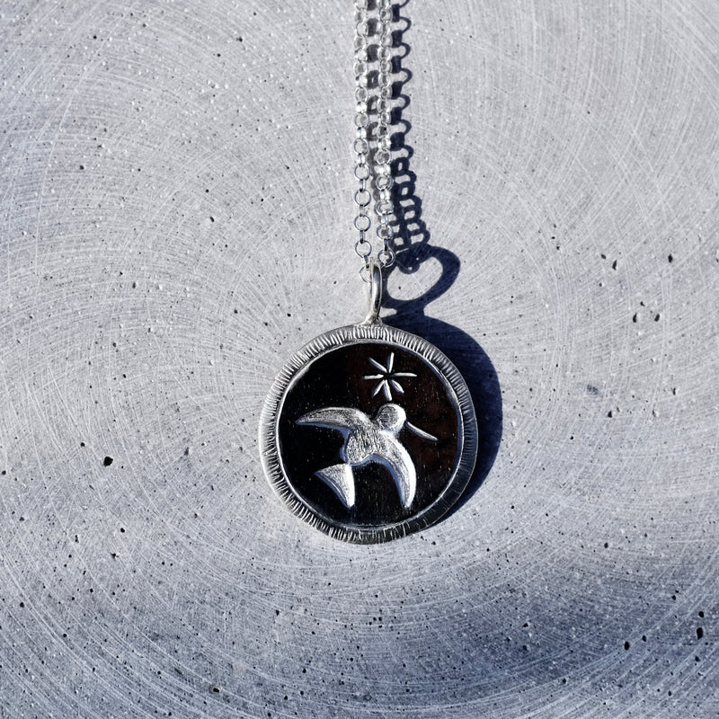 A silver pendant necklace featuring a bird and star design on a dark circular background, resting on a textured gray concrete surface.