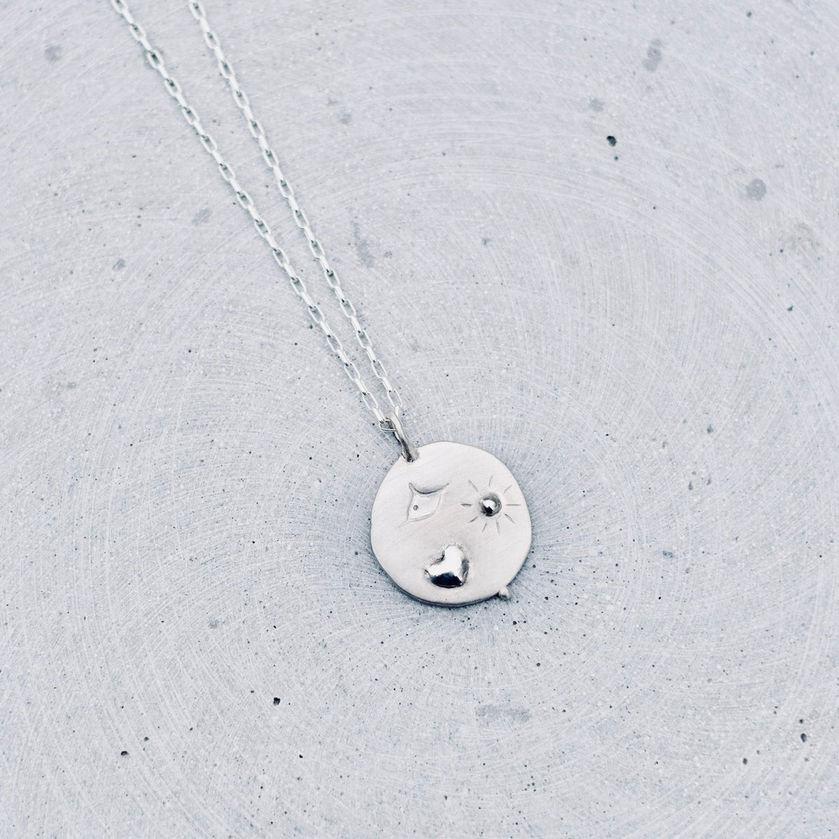 A silver pendant necklace featuring a hand-stamped heart, bird, and radiant sun motif. Hanging from a delicate silver chain against a textured light gray concrete background.