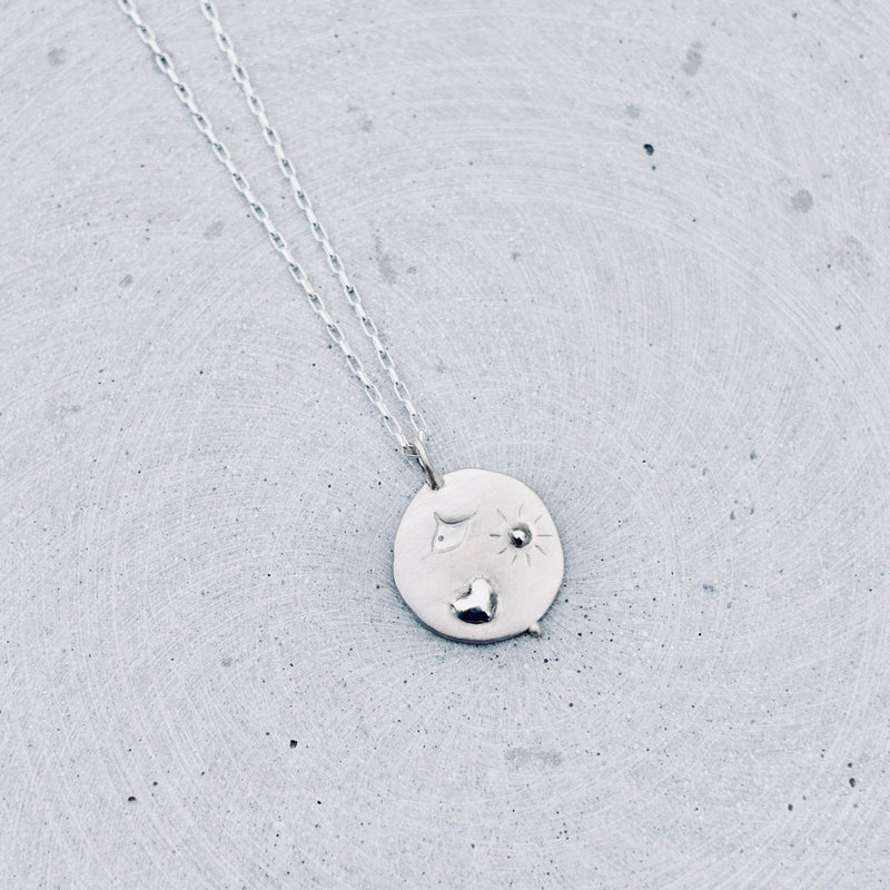 A silver pendant necklace featuring a hand-stamped heart, bird, and radiant sun motif. Hanging from a delicate silver chain against a textured light gray concrete background.