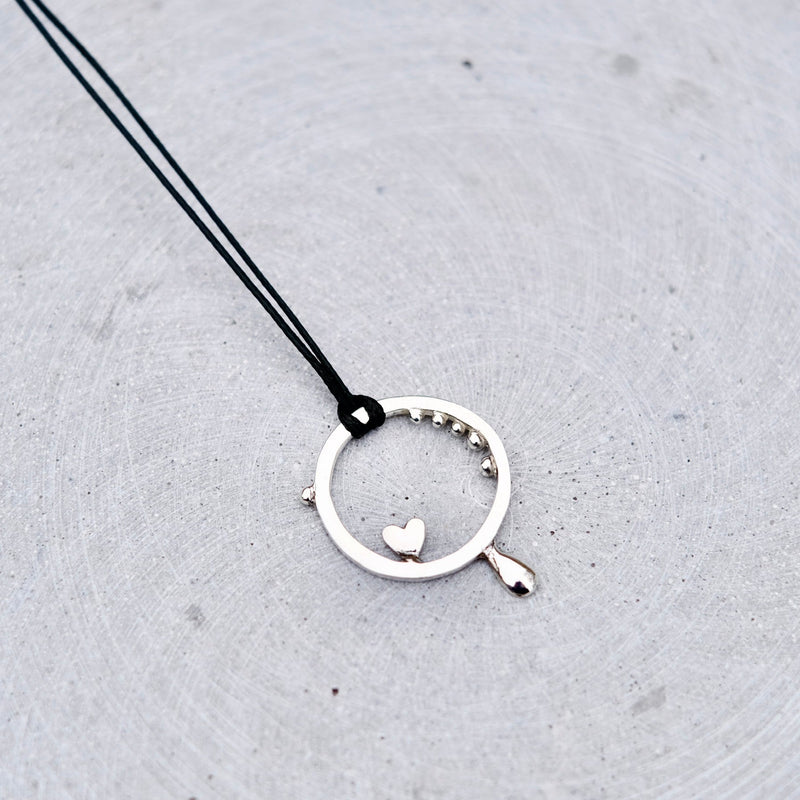 A silver circular pendant necklace with a small dangling heart and a tiny bead detail, suspended on a black cord against a textured gray concrete background.