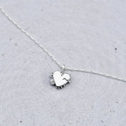 A delicate silver necklace with a small heart-shaped pendant resting on a textured light gray surface.