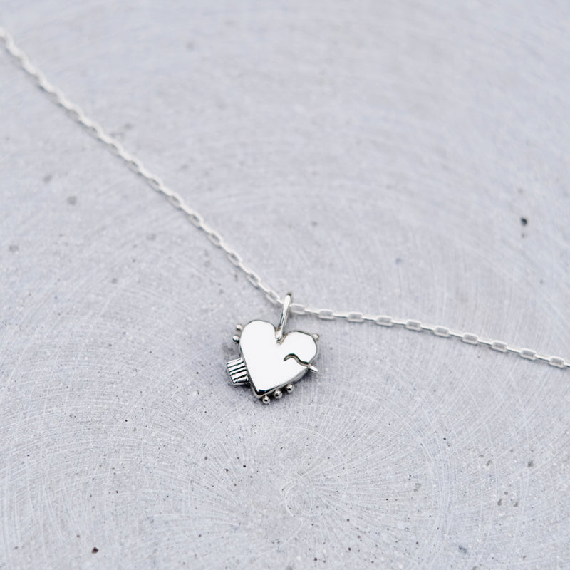 A delicate silver necklace with a small heart-shaped pendant resting on a textured light gray surface.