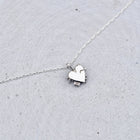 A delicate silver chain necklace featuring a small, polished silver heart-shaped pendant with a tiny dangling charm, displayed on a textured light gray surface.