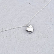 A delicate silver chain necklace featuring a small, polished silver heart-shaped pendant with a tiny dangling charm, displayed on a textured light gray surface.