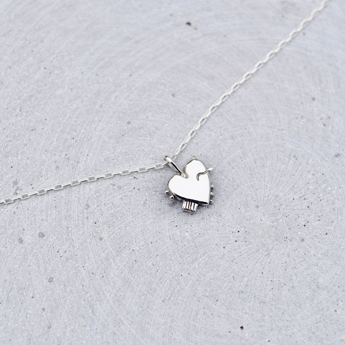 A delicate silver chain necklace featuring a small, polished silver heart-shaped pendant with a tiny dangling charm, displayed on a textured light gray surface.
