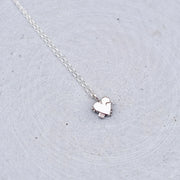 A delicate silver chain necklace with a small, polished silver heart pendant, resting on a textured light gray surface.