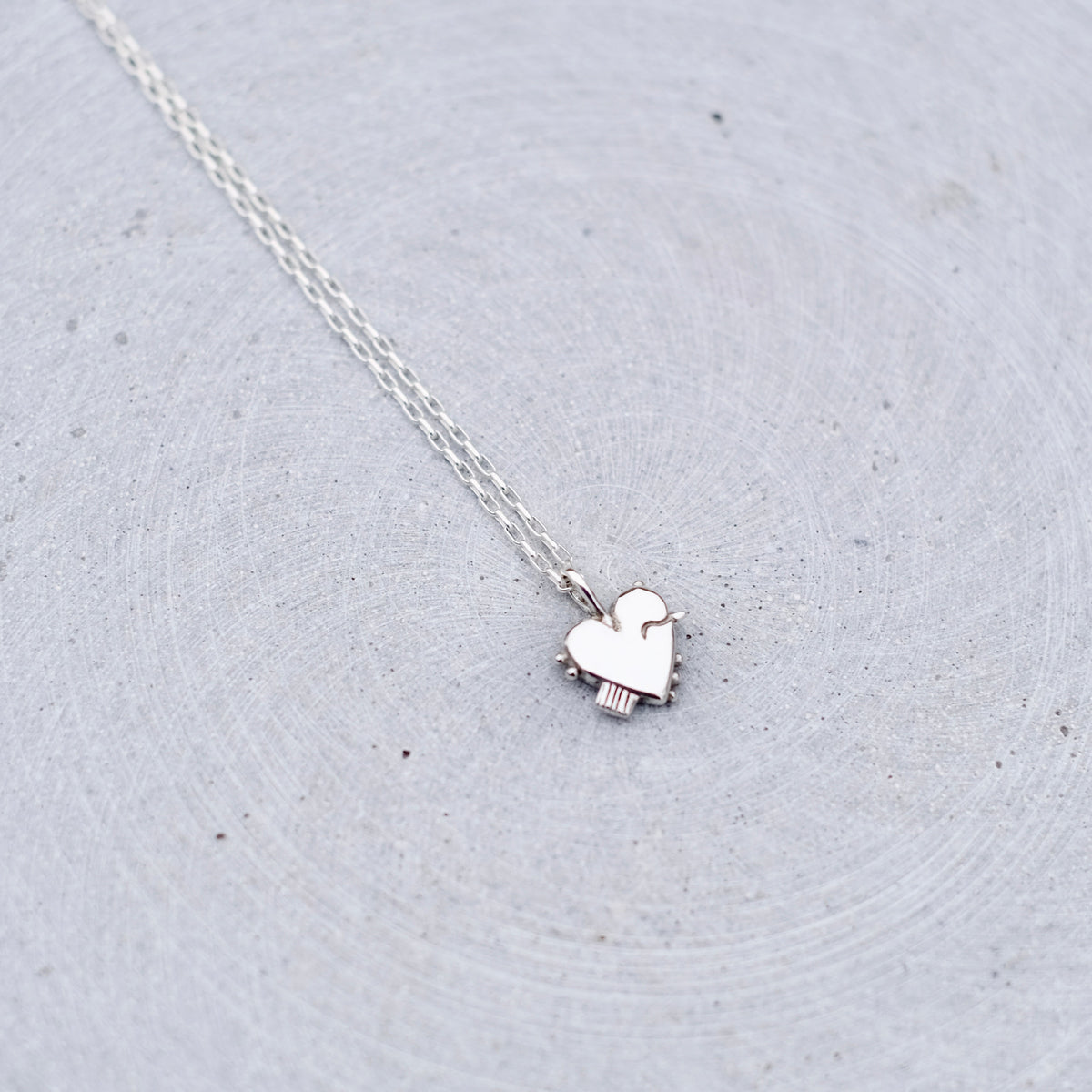 A delicate silver chain necklace with a small, polished silver heart pendant, resting on a textured light gray surface.