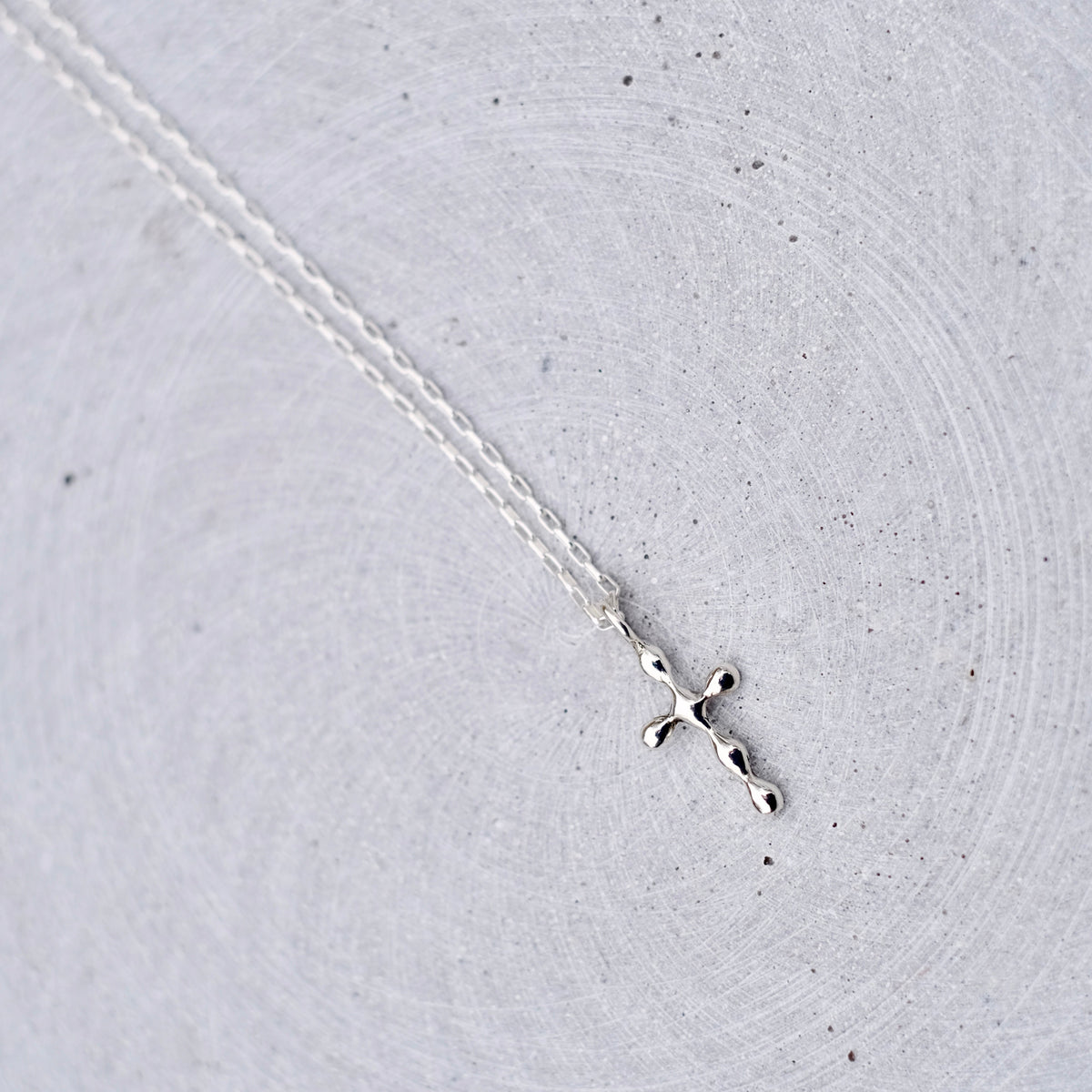 A delicate silver cross pendant necklace on a fine silver chain, displayed on a textured light gray concrete surface.