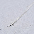 A delicate silver cross pendant necklace on a fine silver chain, displayed on a textured light gray circular surface.