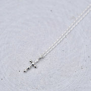 A delicate silver cross pendant necklace on a fine silver chain, displayed on a textured light gray circular surface.