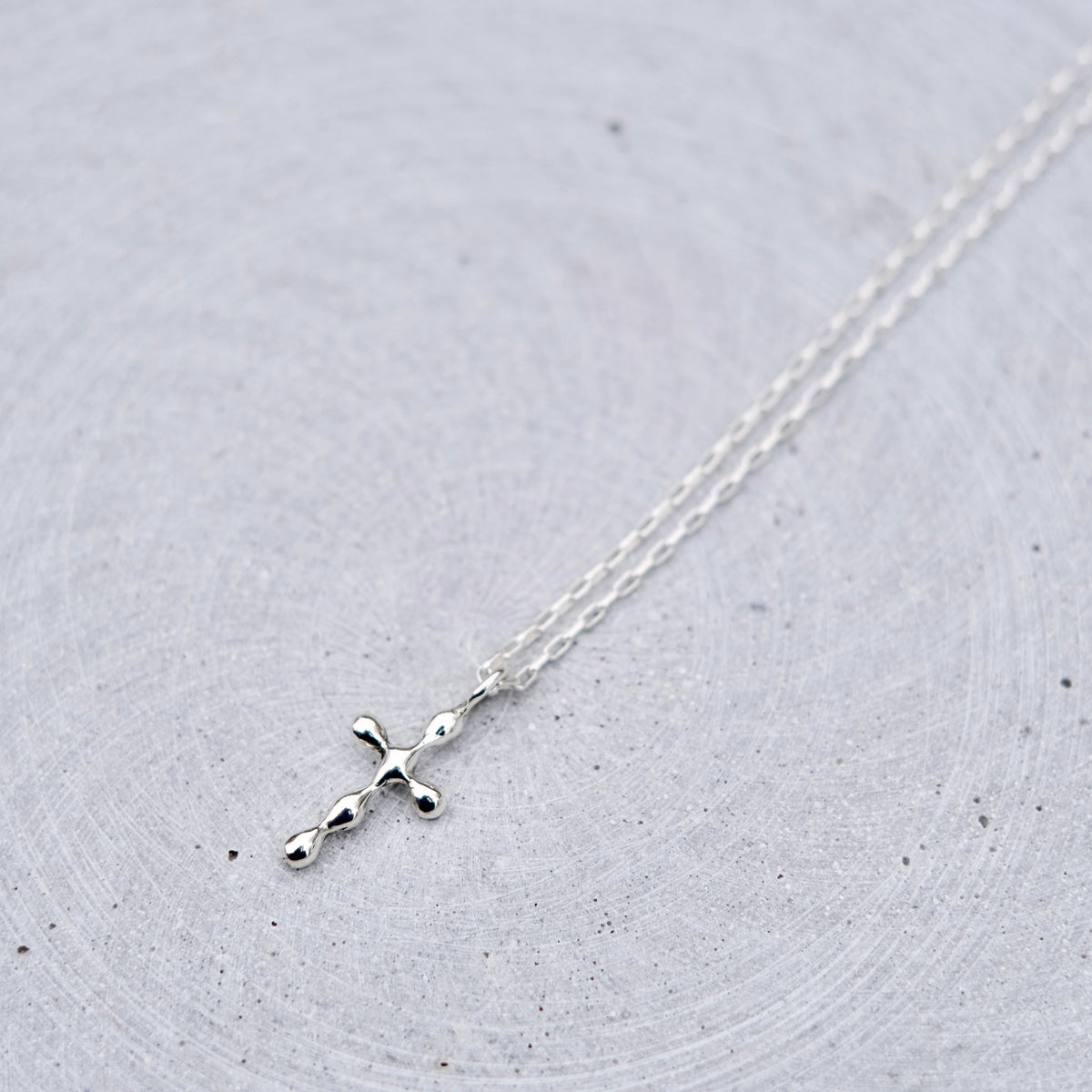 A delicate silver cross pendant necklace on a fine silver chain, displayed on a textured light gray circular surface.