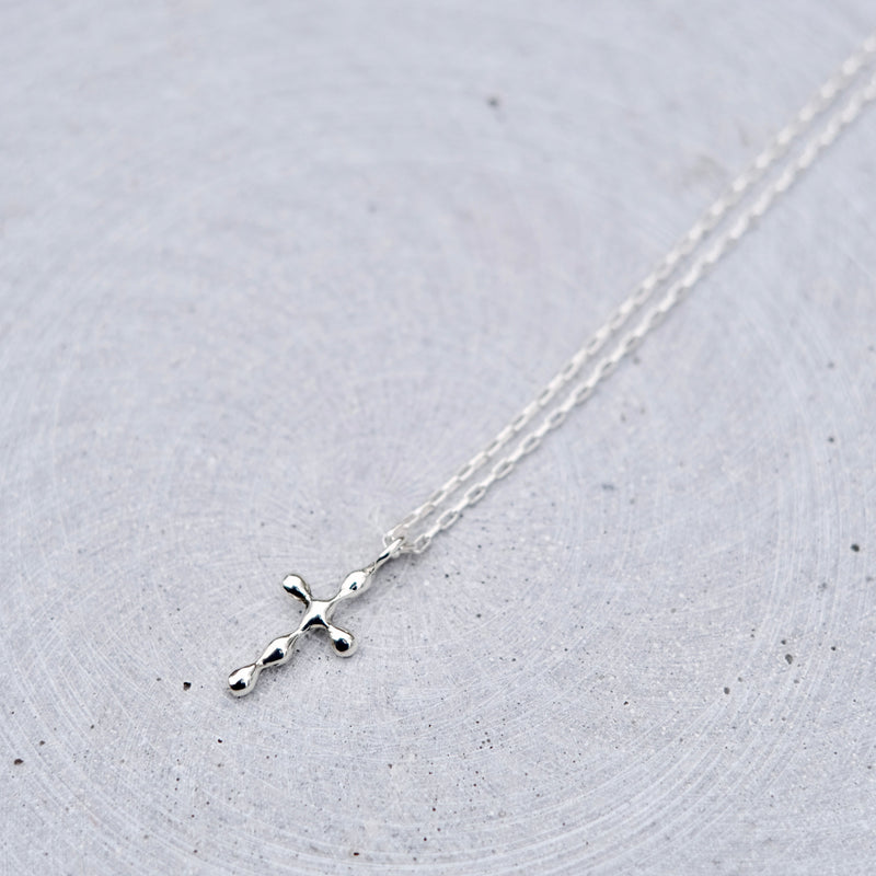 A delicate silver cross pendant necklace on a fine silver chain, displayed on a textured light gray circular surface.