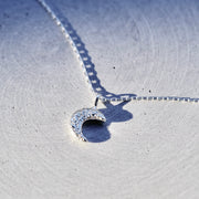 A silver crescent moon pendant necklace on a matching chain, resting on a textured light gray surface with a sharp shadow.