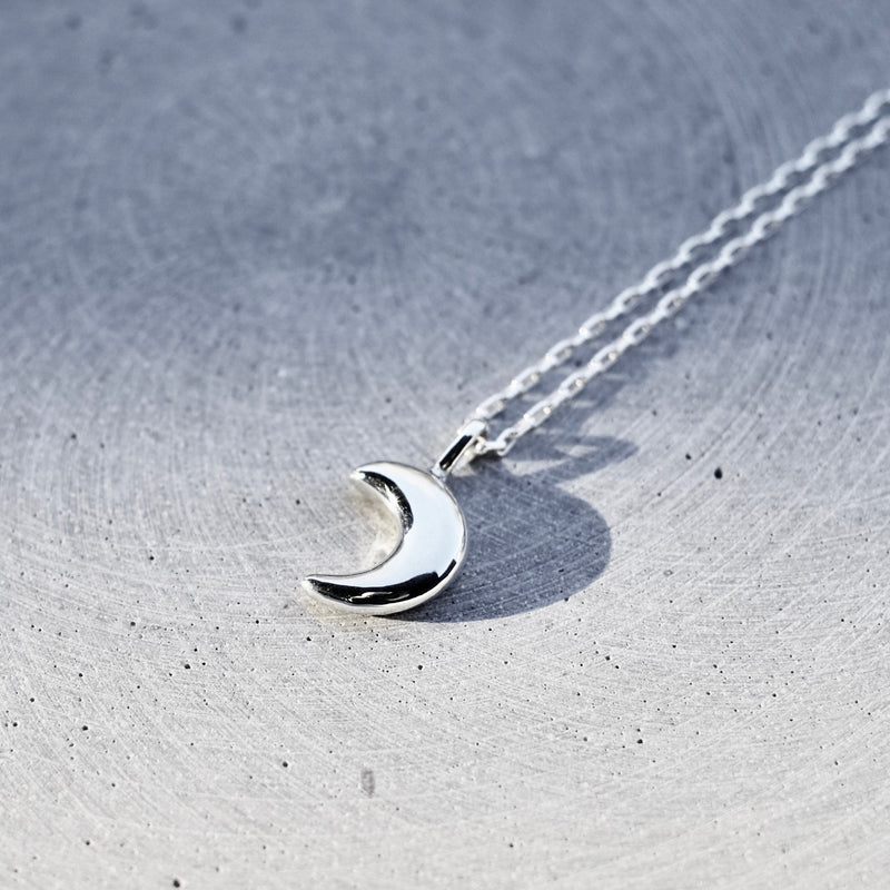 A silver crescent moon pendant necklace on a delicate silver chain, resting on a textured gray stone surface.