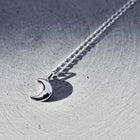 A silver crescent moon pendant necklace on a matching chain, displayed on a textured gray metallic surface.