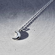 A silver crescent moon pendant necklace on a matching chain, displayed on a textured gray metallic surface.