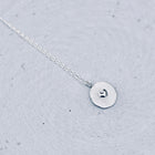 A delicate silver necklace with a small, flat circular disc pendant featuring a tiny heart engraving, resting on a textured light gray surface.