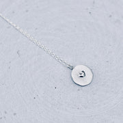 A delicate silver necklace with a small, flat circular disc pendant featuring a tiny heart engraving, resting on a textured light gray surface.