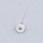 A silver necklace with a textured circular pendant featuring a small heart design, resting on a light gray concrete surface.