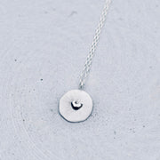 A silver necklace with a textured circular pendant featuring a small heart design, resting on a light gray concrete surface.