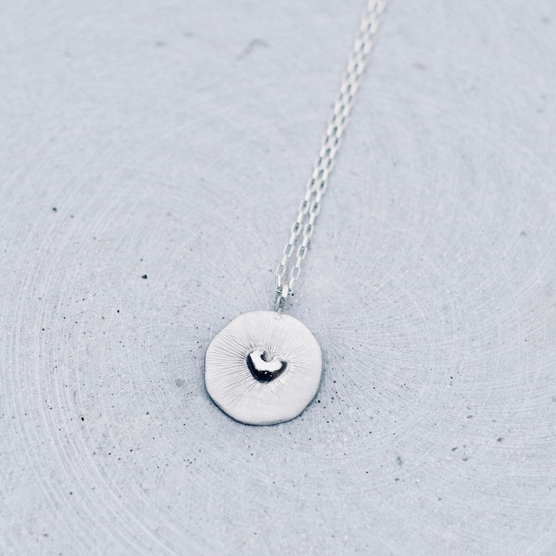 A silver necklace with a textured circular pendant featuring a small heart design, resting on a light gray concrete surface.
