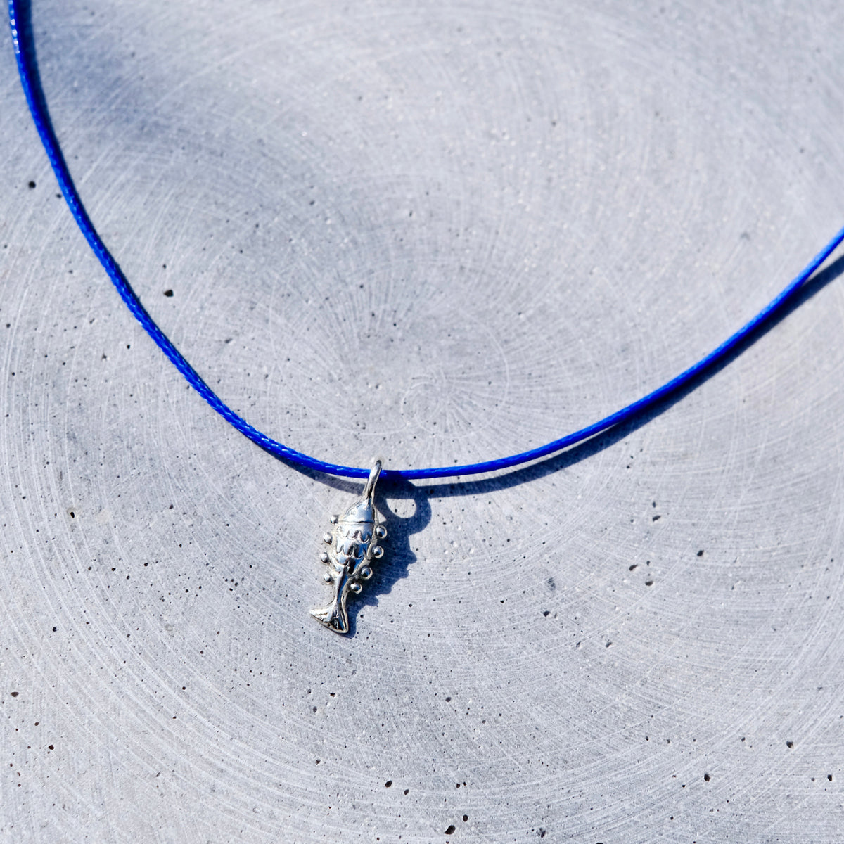 A silver fish pendant necklace on a vibrant blue cord, laid flat on a textured light gray concrete surface.