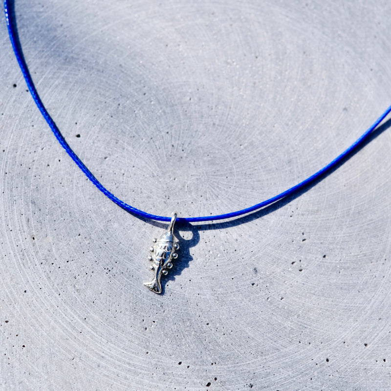 A silver fish pendant necklace on a vibrant blue cord, laid flat on a textured light gray concrete surface.