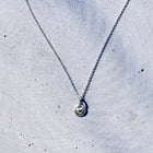 A delicate silver chain necklace with a small, round, textured silver pendant, laid out on a light gray concrete surface.