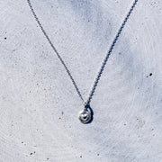 A delicate silver chain necklace with a small, round, textured silver pendant, laid out on a light gray concrete surface.