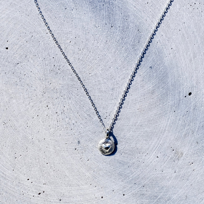 A delicate silver chain necklace with a small, round, textured silver pendant, laid out on a light gray concrete surface.