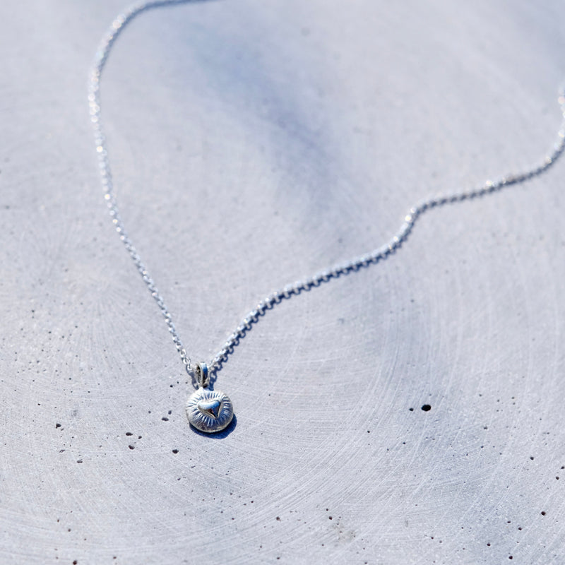 A delicate silver chain necklace with a small, round, textured silver pendant, laid out on a light gray concrete surface.