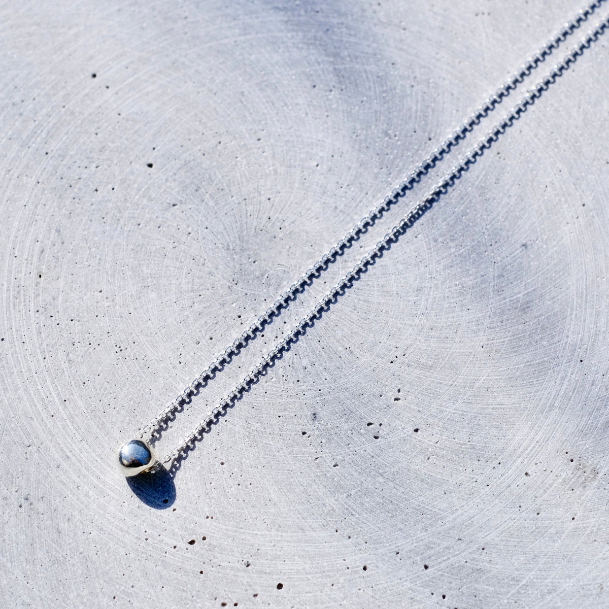 A delicate silver chain necklace with a small gold-toned ring setting holding a round, iridescent blue-gray bead pendant, laid diagonally on a textured light gray concrete surface.