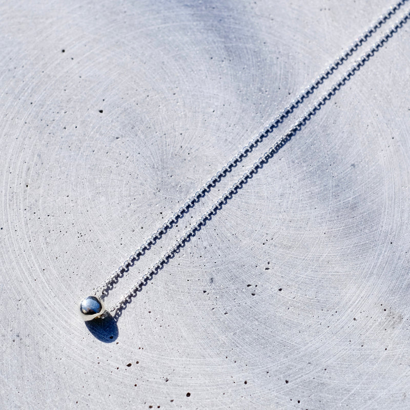 A delicate silver chain necklace with a small gold-toned ring setting holding a round, iridescent blue-gray bead pendant, laid diagonally on a textured light gray concrete surface.