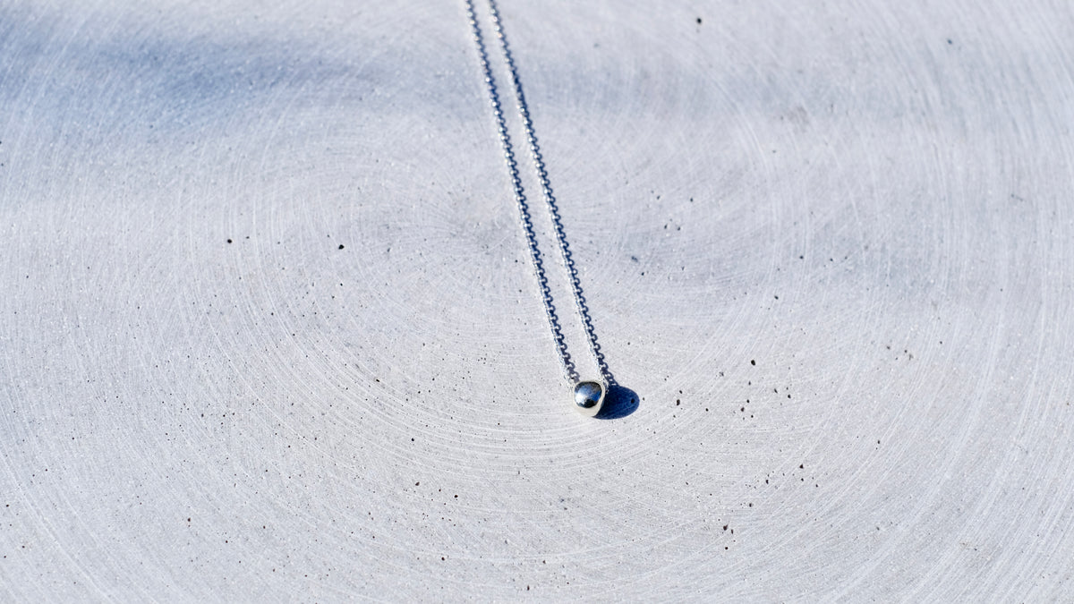 A delicate silver chain necklace with a small, polished silver bead pendant, laid out on a textured, light gray concrete surface.