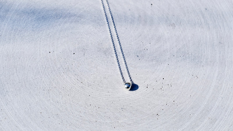A delicate silver chain necklace with a small, polished silver bead pendant, laid out on a textured, light gray concrete surface.