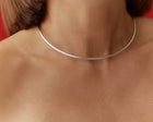 A close-up of a person's neck and upper chest wearing a very thin, polished silver-toned choker necklace against a red background.