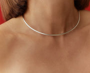 A close-up of a person's neck and upper chest wearing a very thin, polished silver-toned choker necklace against a red background.