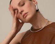 A close-up lifestyle shot of a model wearing a polished silver-toned metal collar necklace and matching geometric drop earrings against a solid warm beige background.