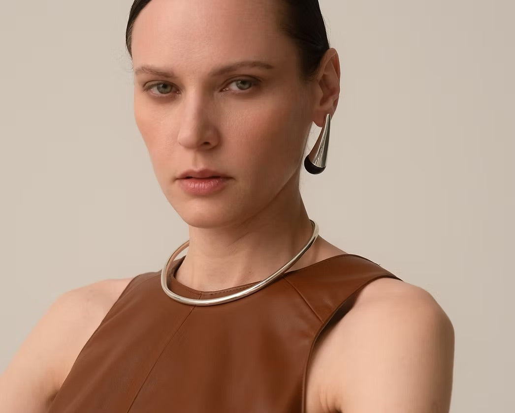 A close-up portrait of a model wearing a minimalist silver-toned metal collar necklace and a matching teardrop-shaped earring, paired with a brown leather-look sleeveless top against a solid light beige background.