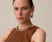 A close-up portrait of a model wearing a minimalist silver-toned metal collar necklace and a matching teardrop-shaped earring, paired with a brown leather-look sleeveless top against a solid light beige background.