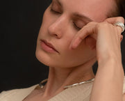A close-up lifestyle shot of a woman with her eyes closed, resting her head on her hand, wearing a chunky silver ring and a silver collar necklace. She is wearing a cream-colored knit top against a dark, solid background.