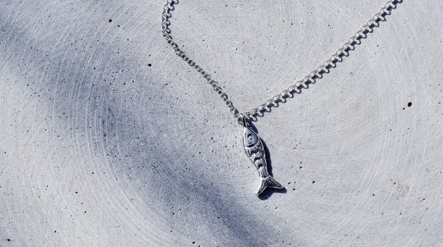 A silver fish pendant necklace on a delicate silver chain, displayed on a textured gray concrete surface.