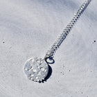 A silver pendant necklace with a textured, hammered circular disc pendant, resting on a light gray concrete surface.