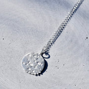 A silver pendant necklace with a textured, hammered circular disc pendant, resting on a light gray concrete surface.
