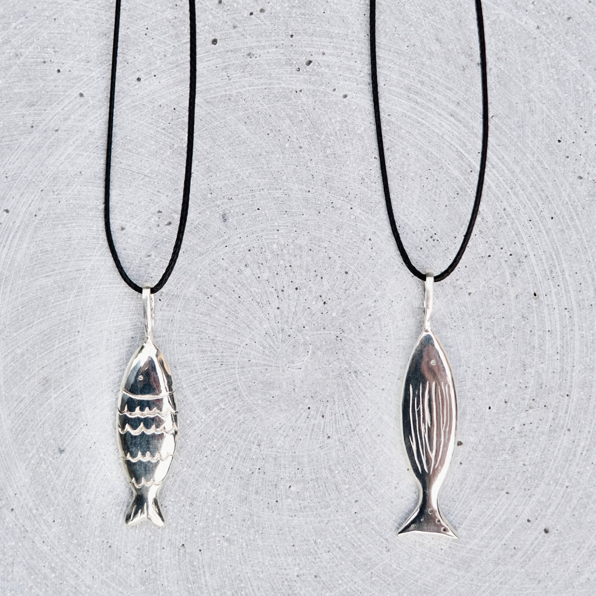Two silver fish-shaped pendant necklaces on black cords, displayed against a textured light gray background.